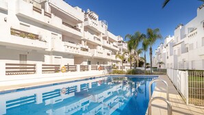 A swimming pool adjacent to a white building with balconies, palm trees, and a clear blue sky. - Valle Romano Prestigious 2266 (Estepona)
