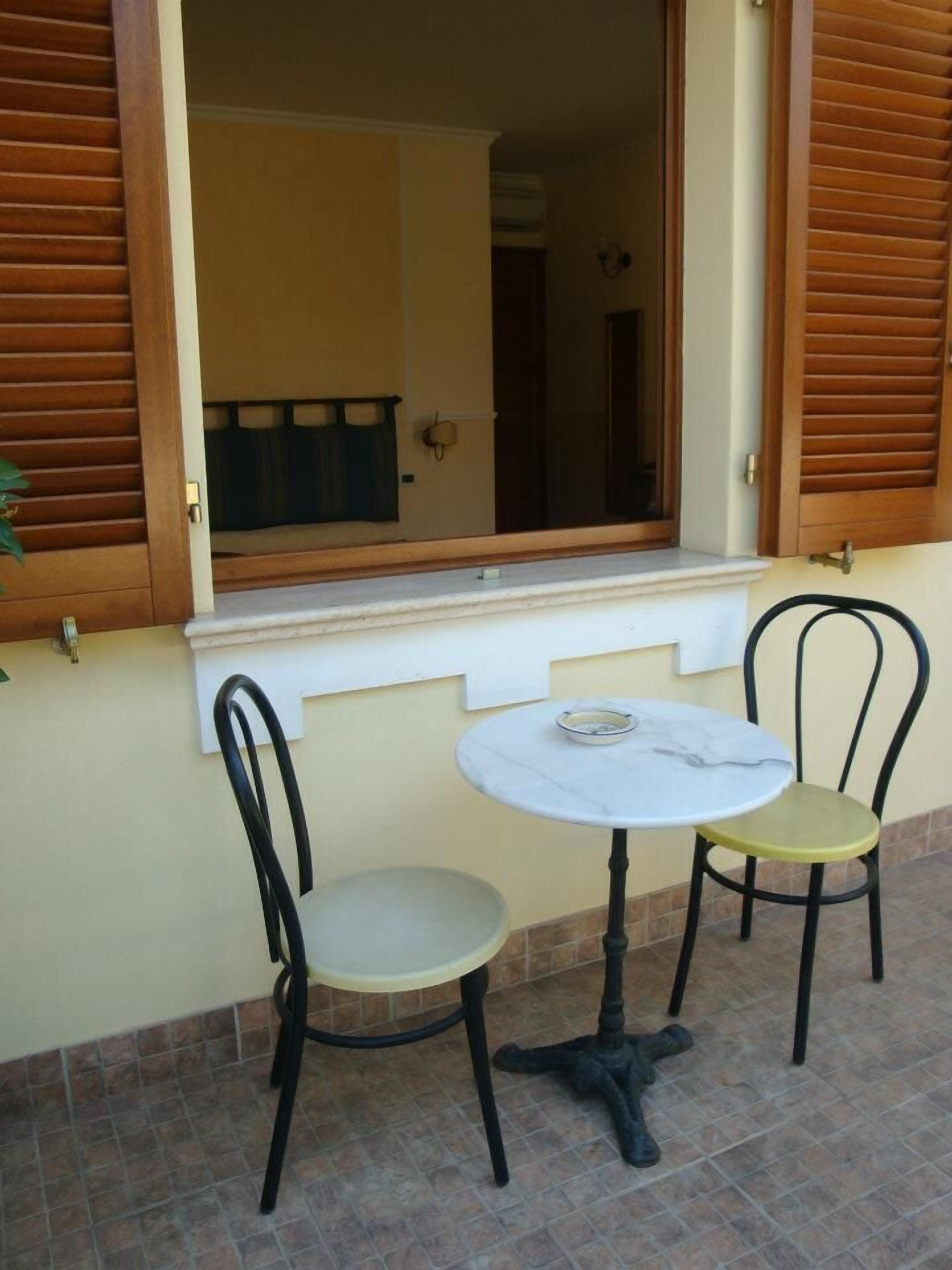 A small outdoor seating area with a round table and two chairs, a window with wooden shutters, and a tiled floor.