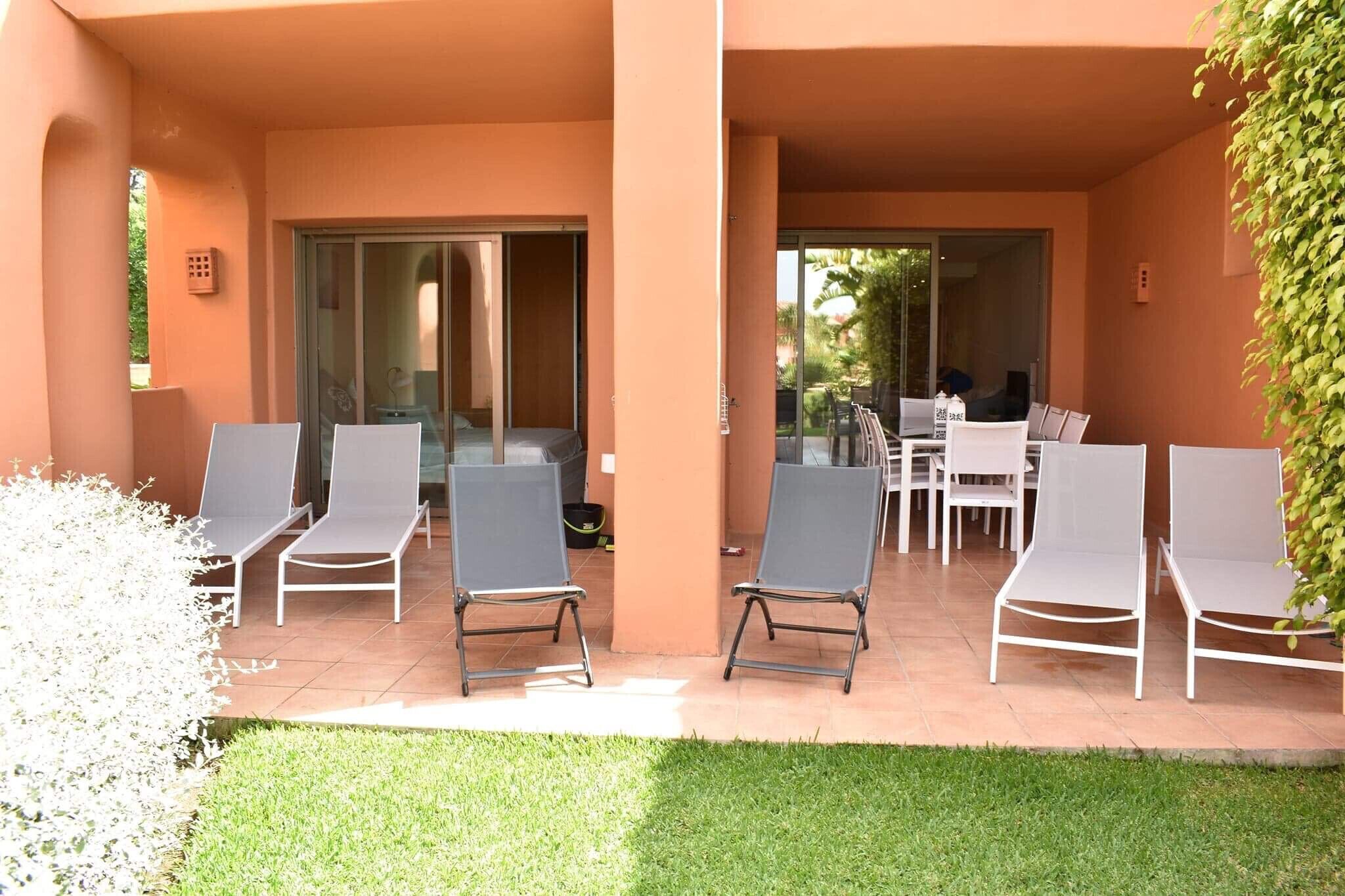 Un área de estar al aire libre cubierta con sillones reclinables y una mesa, rodeada de un jardín y un edificio con puertas correderas de vidrio.