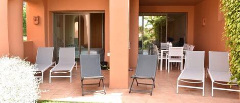 Un área de estar al aire libre cubierta con sillones reclinables y una mesa, rodeada de un jardín y un edificio con puertas correderas de vidrio.