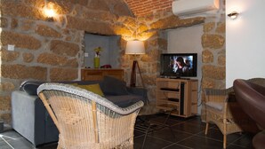 Ein gemütliches Wohnzimmer mit Steinwänden, einem grauen Sofa, Korbstühlen, einem Fernseher und einer Lampe.