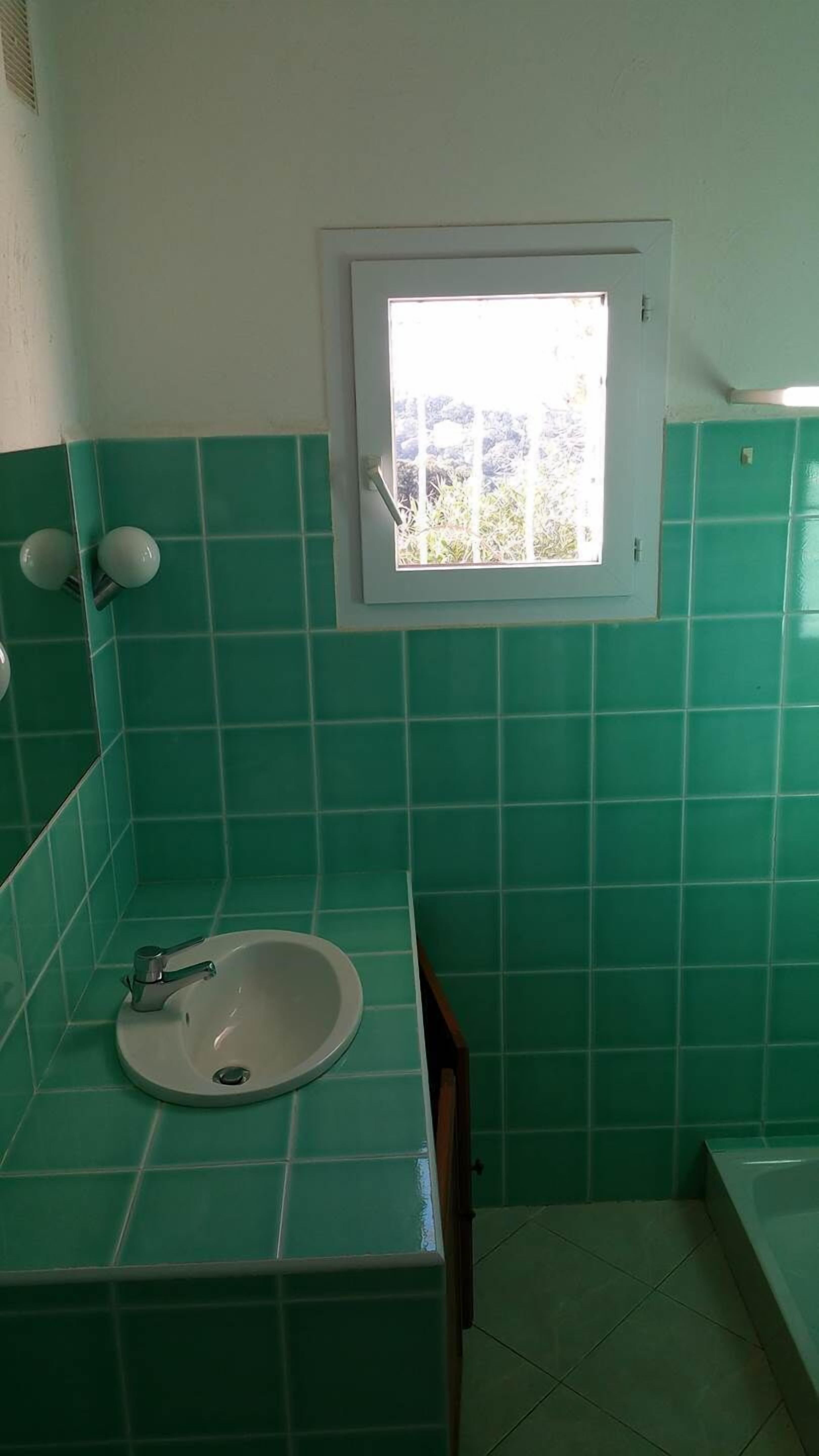Een badkamer met groene tegels, een witte wastafel en een raam boven de spiegel.