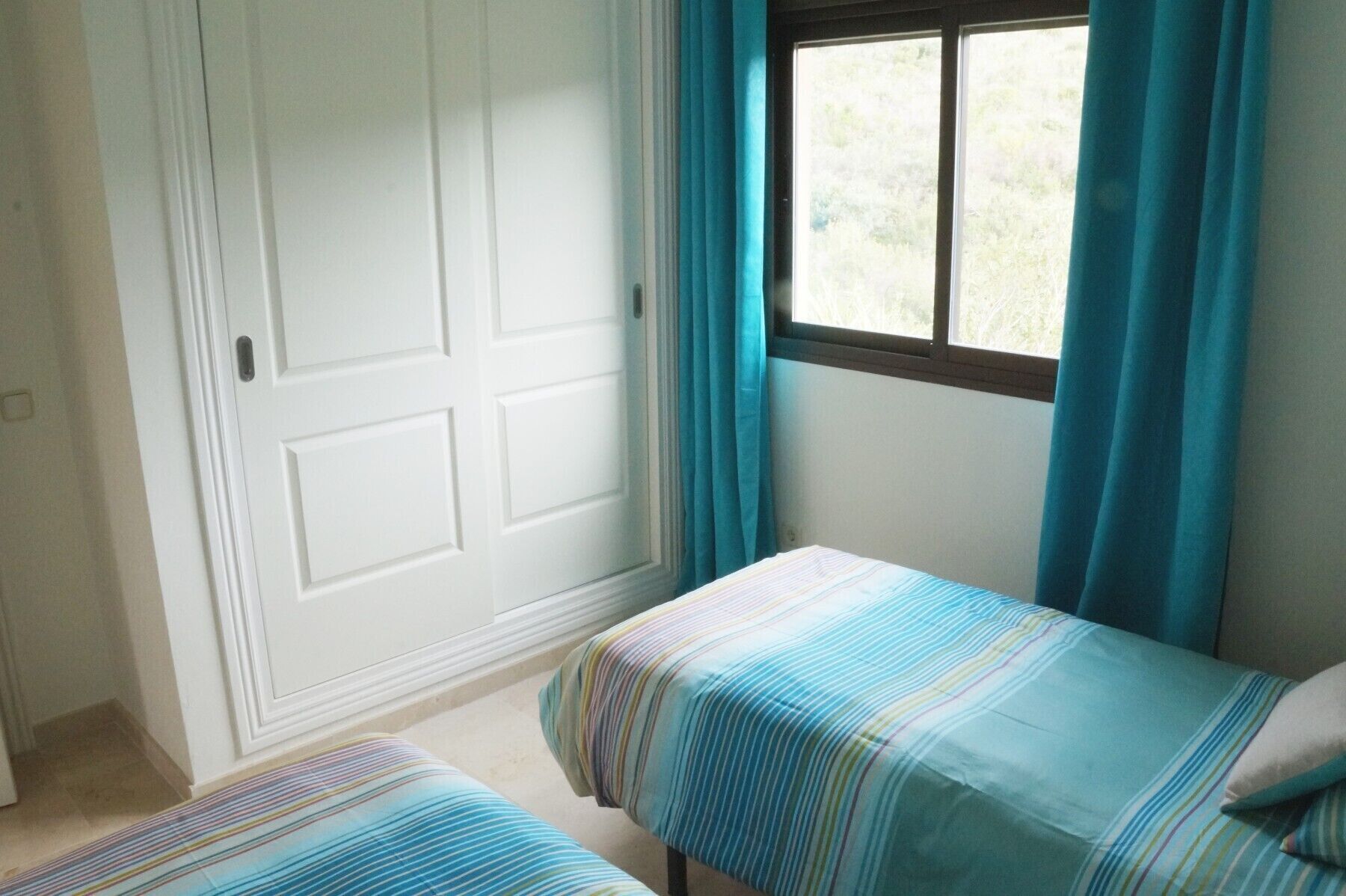 A bedroom with two single beds, a window with curtains, and a white door.