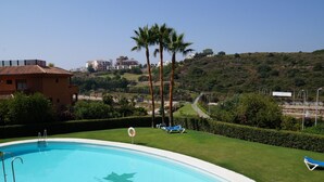 A swimming pool with a palm tree and lounge chairs, surrounded by well-maintained grass and a view of buildings in the background.
