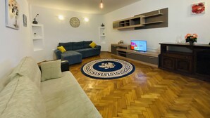 Un soggiorno con un divano, una poltrona blu, un pavimento in parquet a spina di pesce, un tappeto rotondo, una televisione ed scaffali a muro.