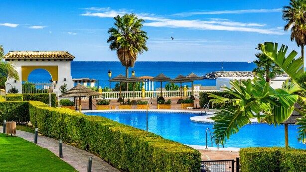 A pool area with a clear view of the ocean, a palm tree, and a building with a yellow archway. - Marina Duquesa 2110 Lovely Place (Manilva)