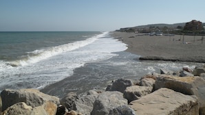 Een strand met een rotsachtige voorgrond, een zandstrand en een gebouw op de achtergrond.