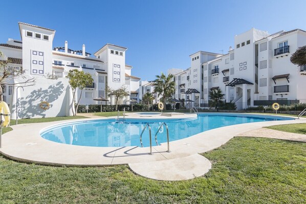 A pool area with a clear blue pool, surrounded by white buildings with balconies and a grassy lawn.