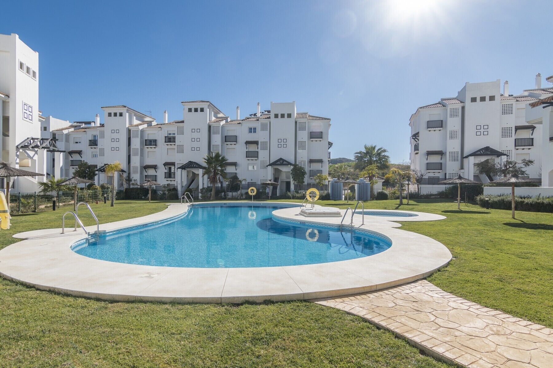A swimming pool with a circular design, surrounded by a well-maintained lawn and a building complex in the background.