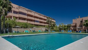 A swimming pool with clear blue water, surrounded by lounge chairs and palm trees, with a multi-story building in the background.