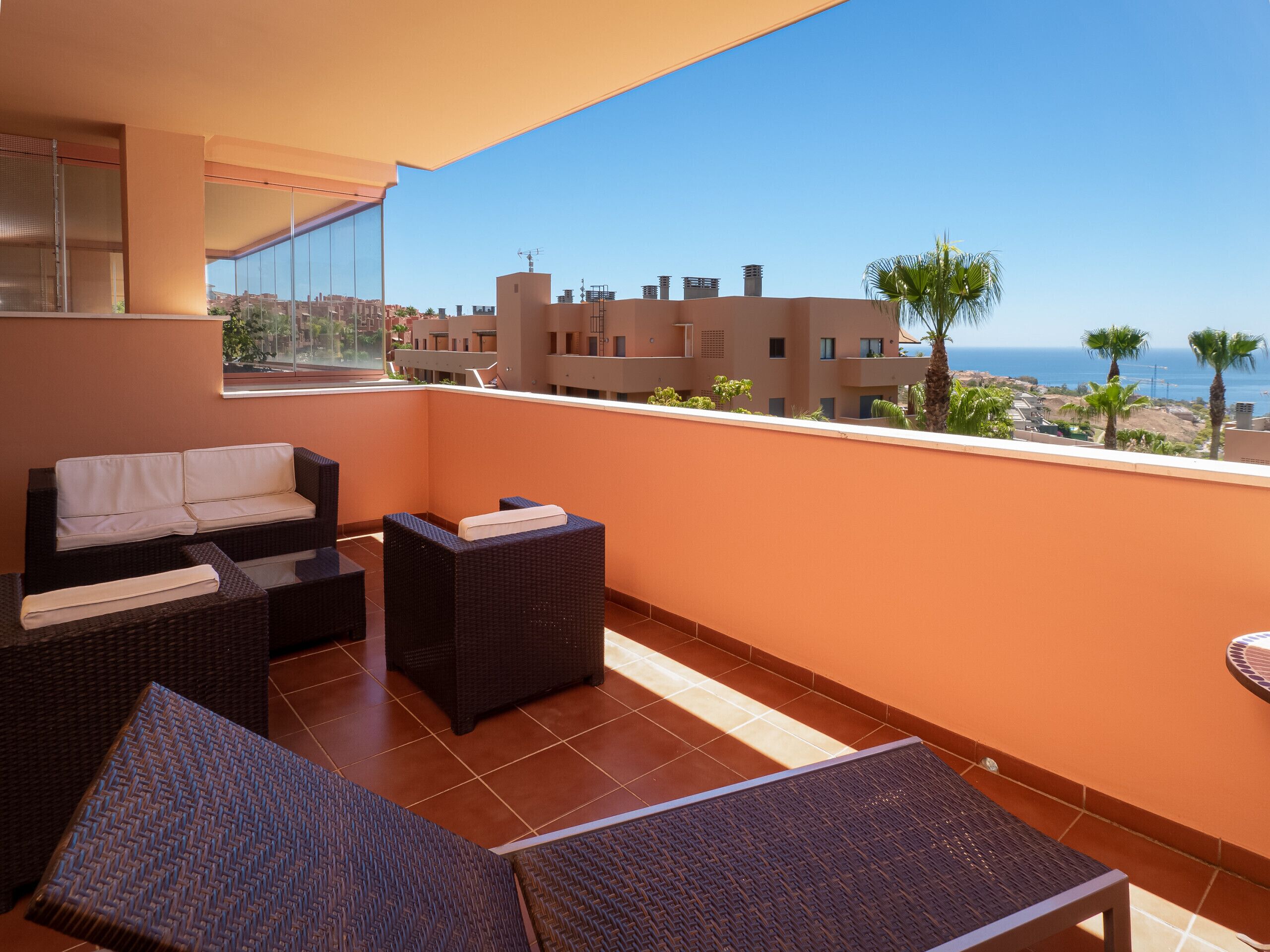 A balcony with wicker furniture, a sofa, and a view of the sea and buildings.