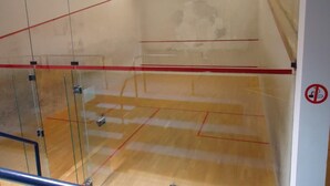 A squash court with glass walls and a wooden floor, marked with red lines.