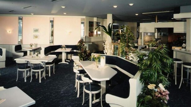 A modern restaurant with white furniture, black cushioned seating, and green plants.