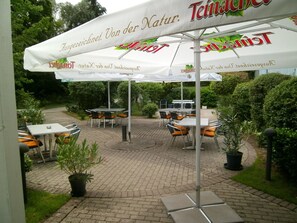 An outdoor seating area with tables and chairs under umbrellas, surrounded by greenery. - Sportpark Jürgen Fassbender (Eggenstein-Leopoldshafen)
