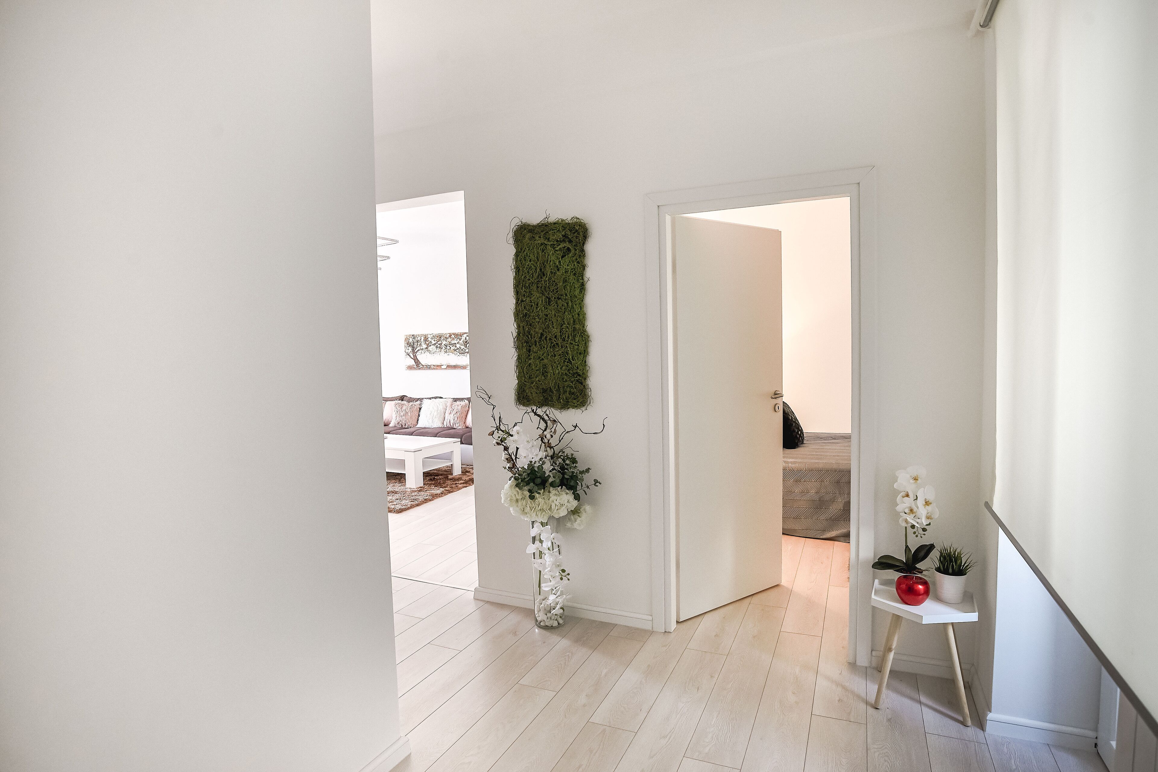 A hallway with a white wall, a green vertical garden, and a small white shelf with a red apple and a potted plant.