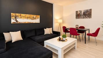 A modern living room with a black sectional sofa, a white coffee table, red chairs, and a dining table. There are two paintings on the walls.