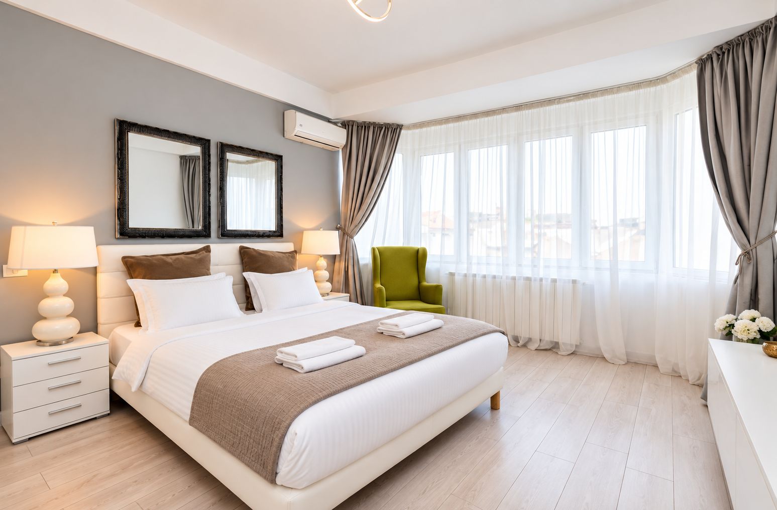 Habitación de hotel con una cama grande, dos mesitas de noche con lámparas, un sillón verde y vistas de la ciudad a través de cortinas translúcidas.
