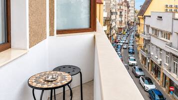 A balcony with a table and two chairs, overlooking a busy street with parked cars and buildings.