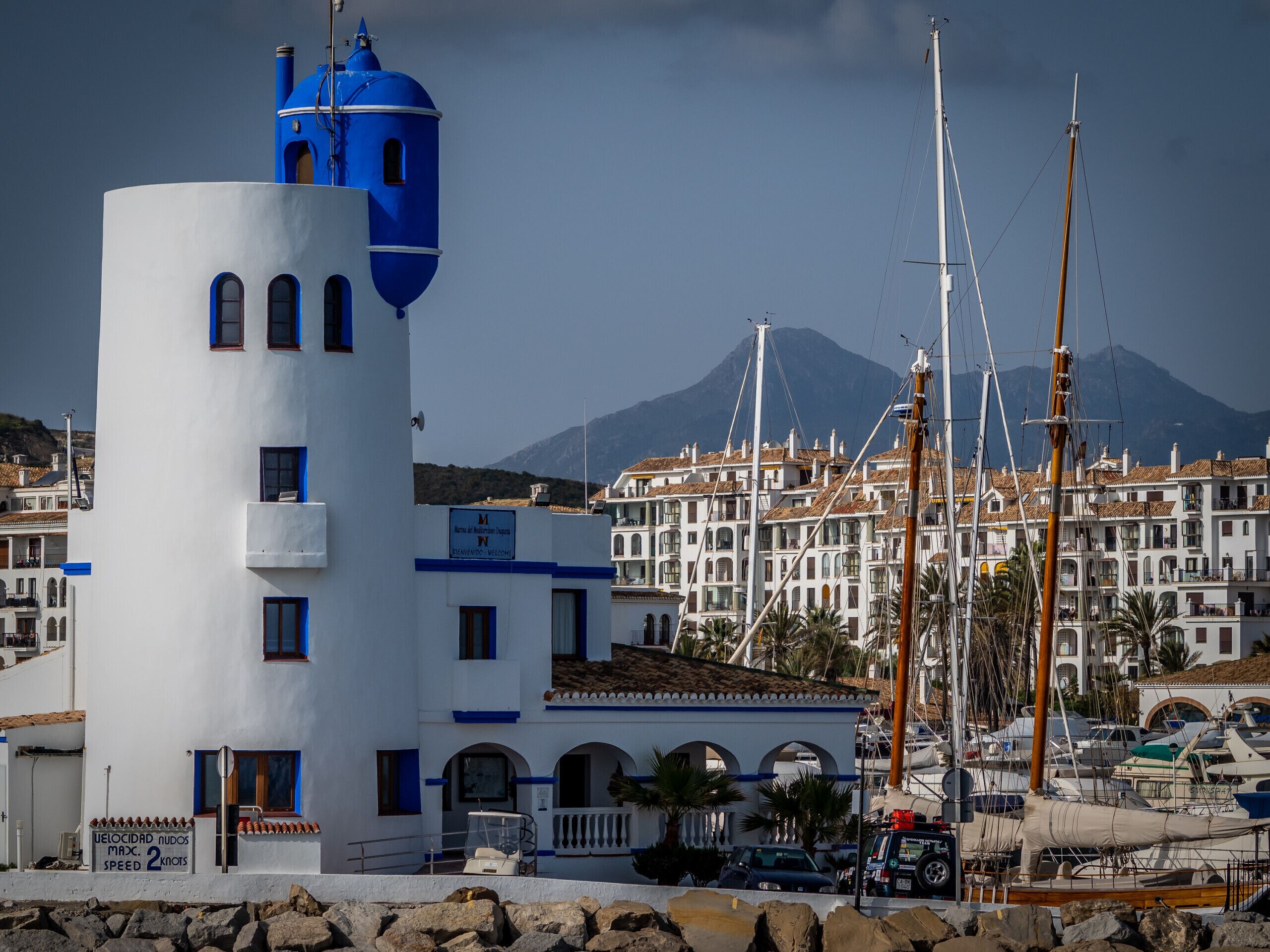 Un bâtiment situé sur la côte, doté d’une tour bleue imposante, un port de plaisance avec des bateaux, et une montagne en arrière-plan.