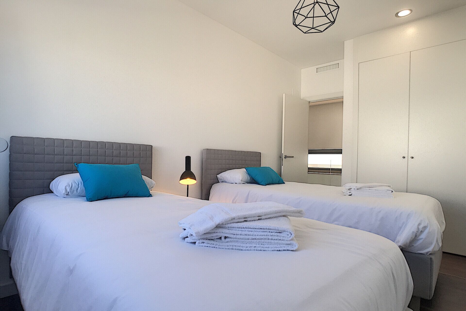 A hotel room with two beds, each with a grey headboard, white bedding, and a teal pillow. There is a lamp on one side and a stack of towels on the other.