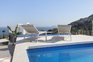 Une piscine avec deux chaises longues et une table, offrant une vue sur un paysage montagneux et la mer.