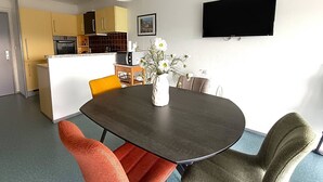 Een moderne woonkamer met een donker houten eettafel, oranje en groene stoelen, een witte vaas met bloemen, een flatscreen televisie aan de muur en een keuken op de achtergrond.