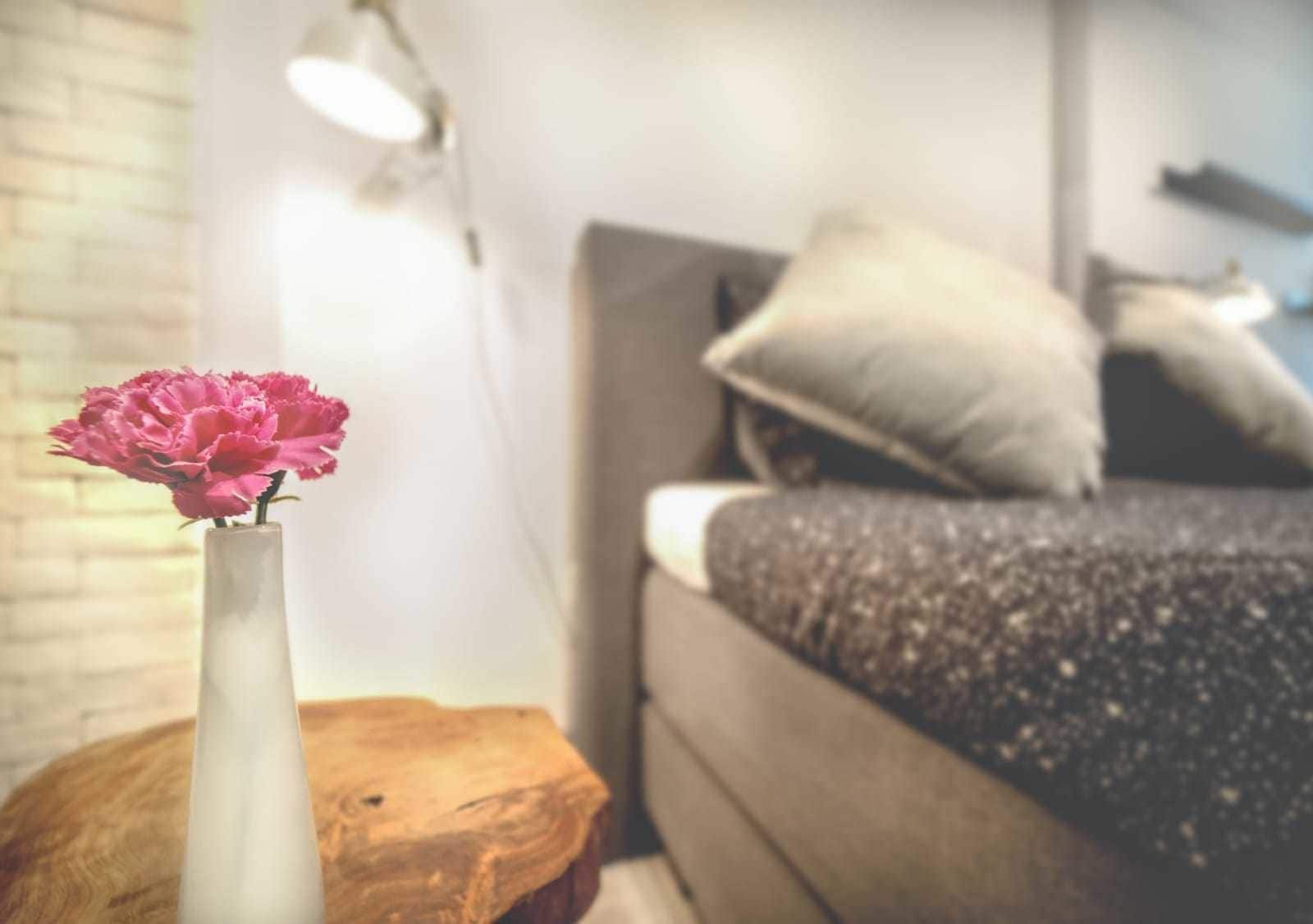Quarto com cama, almofadas, abajur e um vaso de flores sobre um banquinho de madeira.