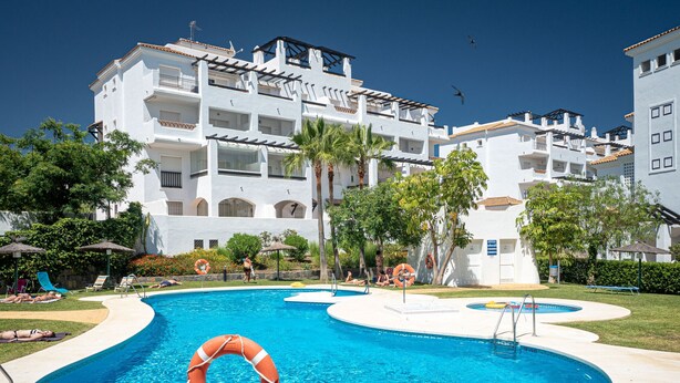 A swimming pool with a lifebuoy, surrounded by lounge chairs and palm trees, with a building in the background. - Residencial Duquesa 2091 Manilva (Manilva)