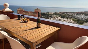 Une table en bois, avec deux verres à vin et une bouteille de vin, placée devant un paysage représentant une ville côtière et la mer.