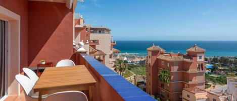 A balcony with a wooden table and white chairs, overlooking a coastal town with buildings and the sea.