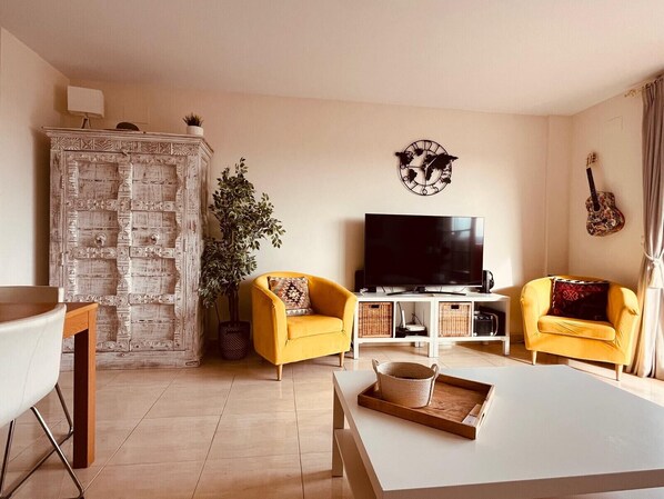 A living room with two yellow armchairs, a white coffee table, a large flat-screen TV, a decorative wall clock, and a guitar on the wall. - Duquesa Regent 2173 Sunset (Manilva)