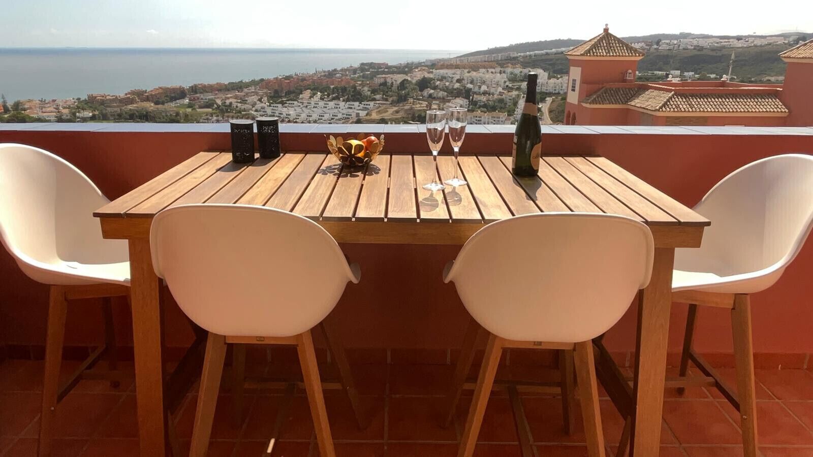 A wooden table with two white chairs on a terrace with a view of a coastal town.