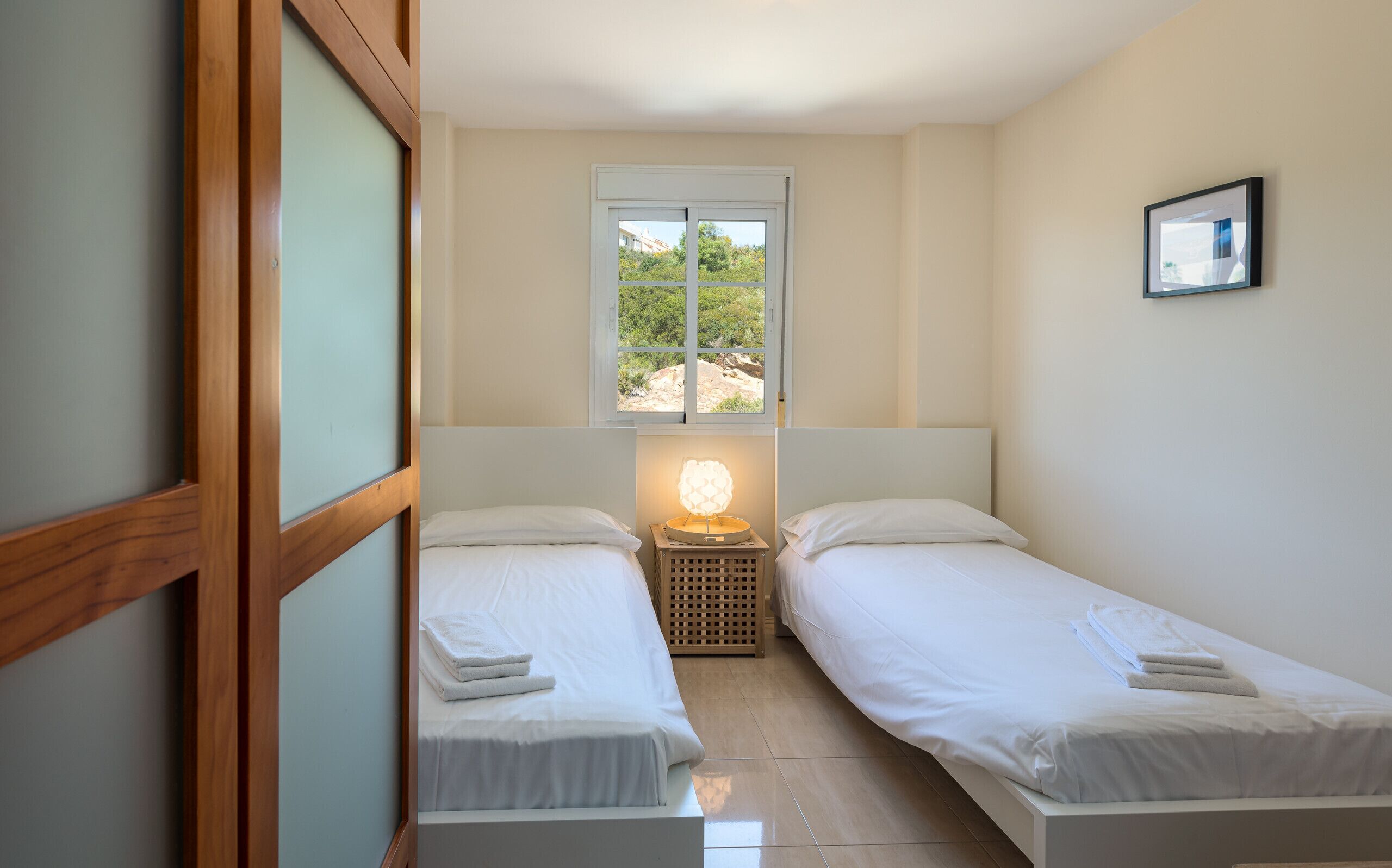 A small, clean room with two single beds, a window with a view of greenery, and a bedside lamp.