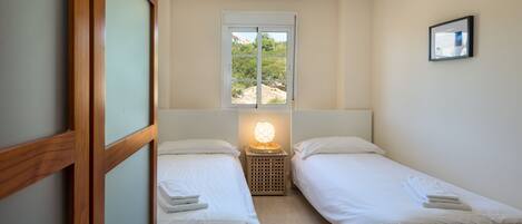 A small, clean room with two single beds, a window with a view of greenery, and a bedside lamp.