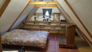 Un pequeño y acogedor dormitorio en ático con techo inclinado, una cama con colcha estampada, un escritorio de madera con libros y objetos decorativos, y una ventana con cortinas.