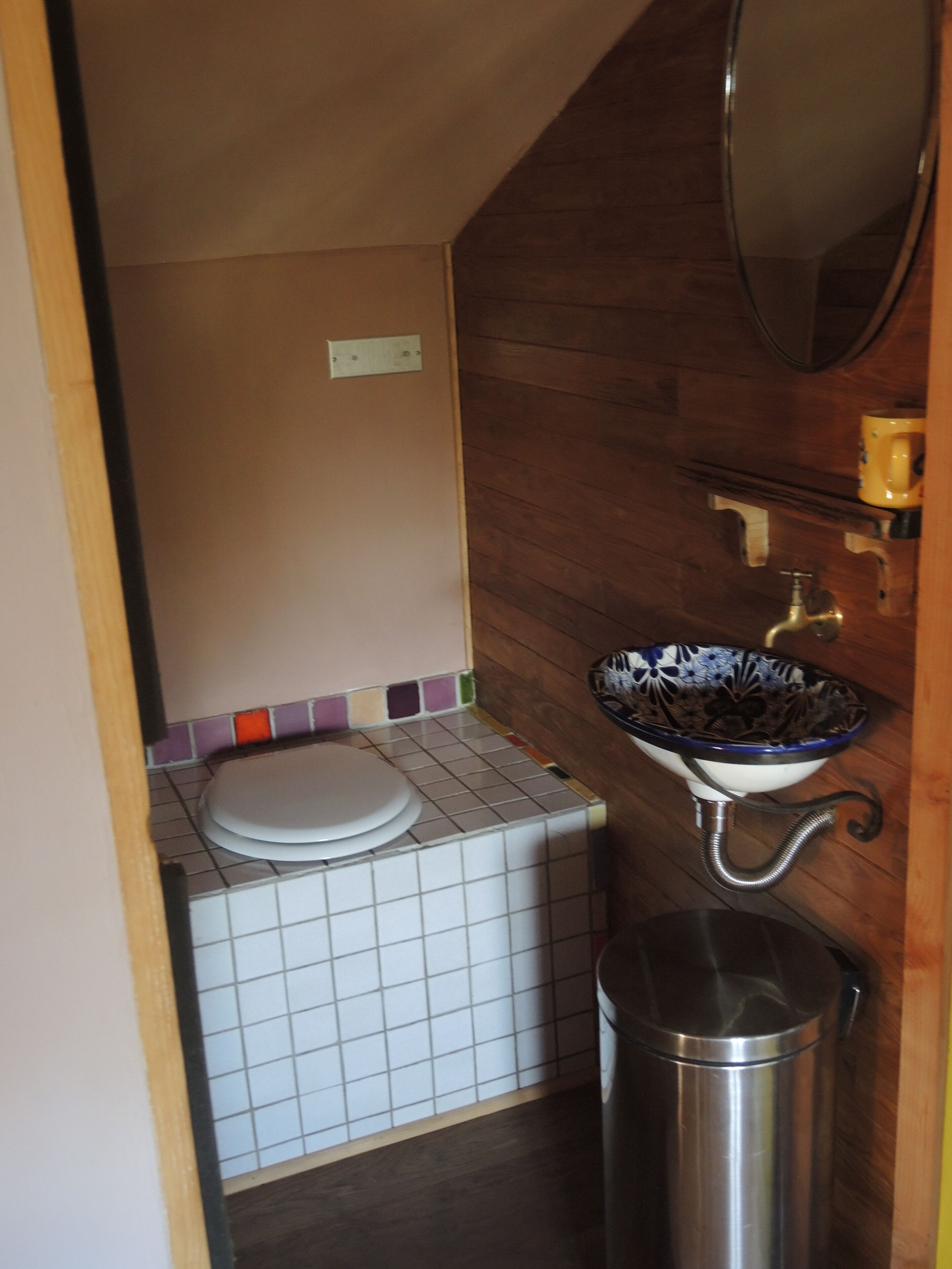 Een compacte badkamer met een wit toilet, een klein wastafeltje met een blauw-witte wasbak en een roestvrijstalen prullenbak.
