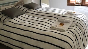 A neatly made bed with striped bedding, a wooden ceiling beam, a large mirror, and a window with blinds.