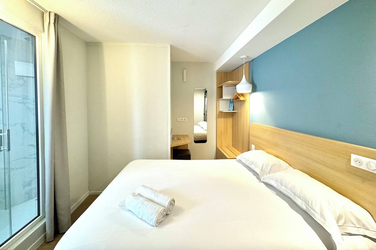 A hotel room with a large bed, a window with curtains, a wooden headboard, and a bedside table with a lamp.