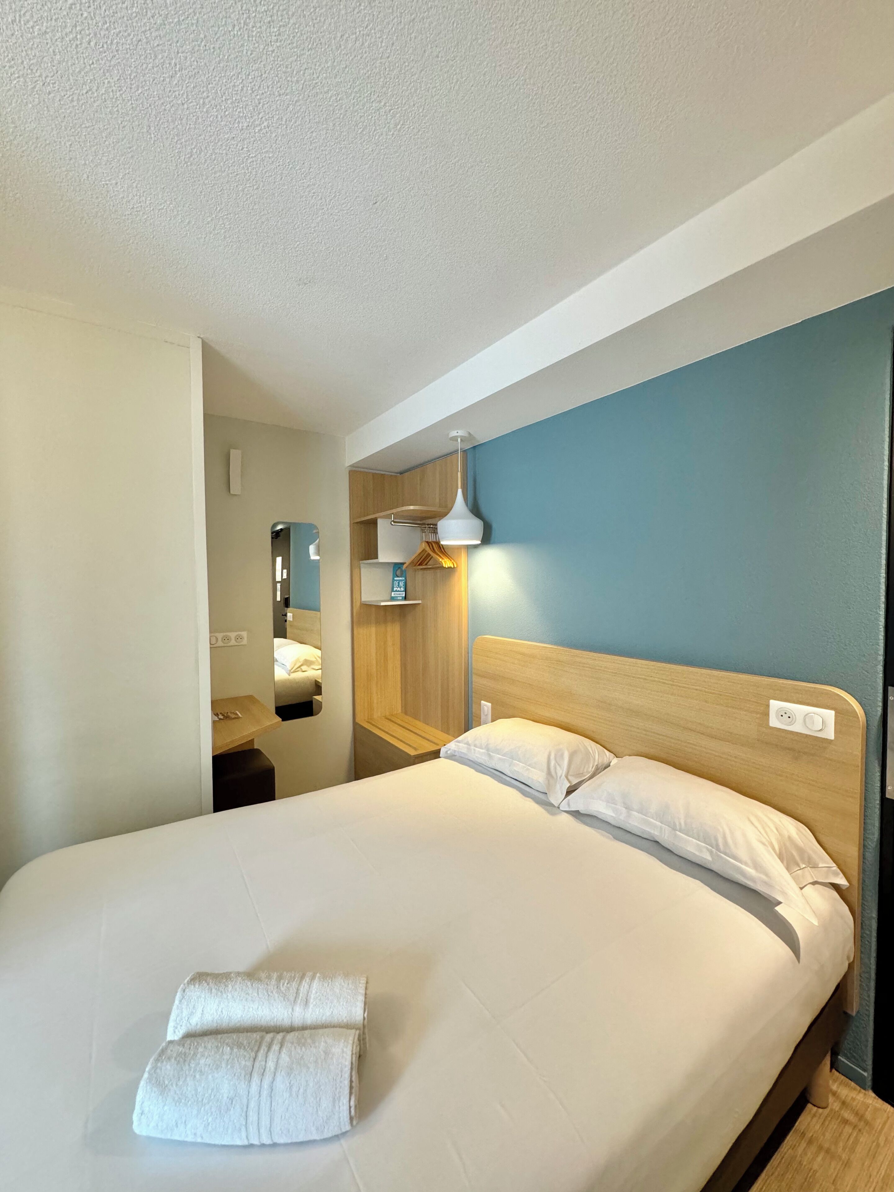 A neatly made bed with white linens and a folded towel, a wooden headboard, a bedside table, a wall-mounted shelf, and a hanging lamp.