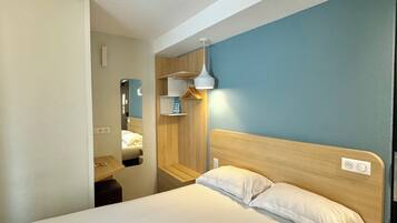 A neatly made bed with white linens and a folded towel, a wooden headboard, a bedside table, a wall-mounted shelf, and a hanging lamp.