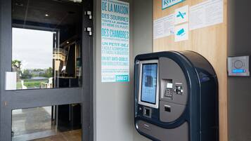 Un kiosque en libre-service doté d’un écran numérique et d’un clavier, une enseigne arborant le logo « Kyriad DIRECT » et une affiche en français décrivant les services offerts par l’hôtel.