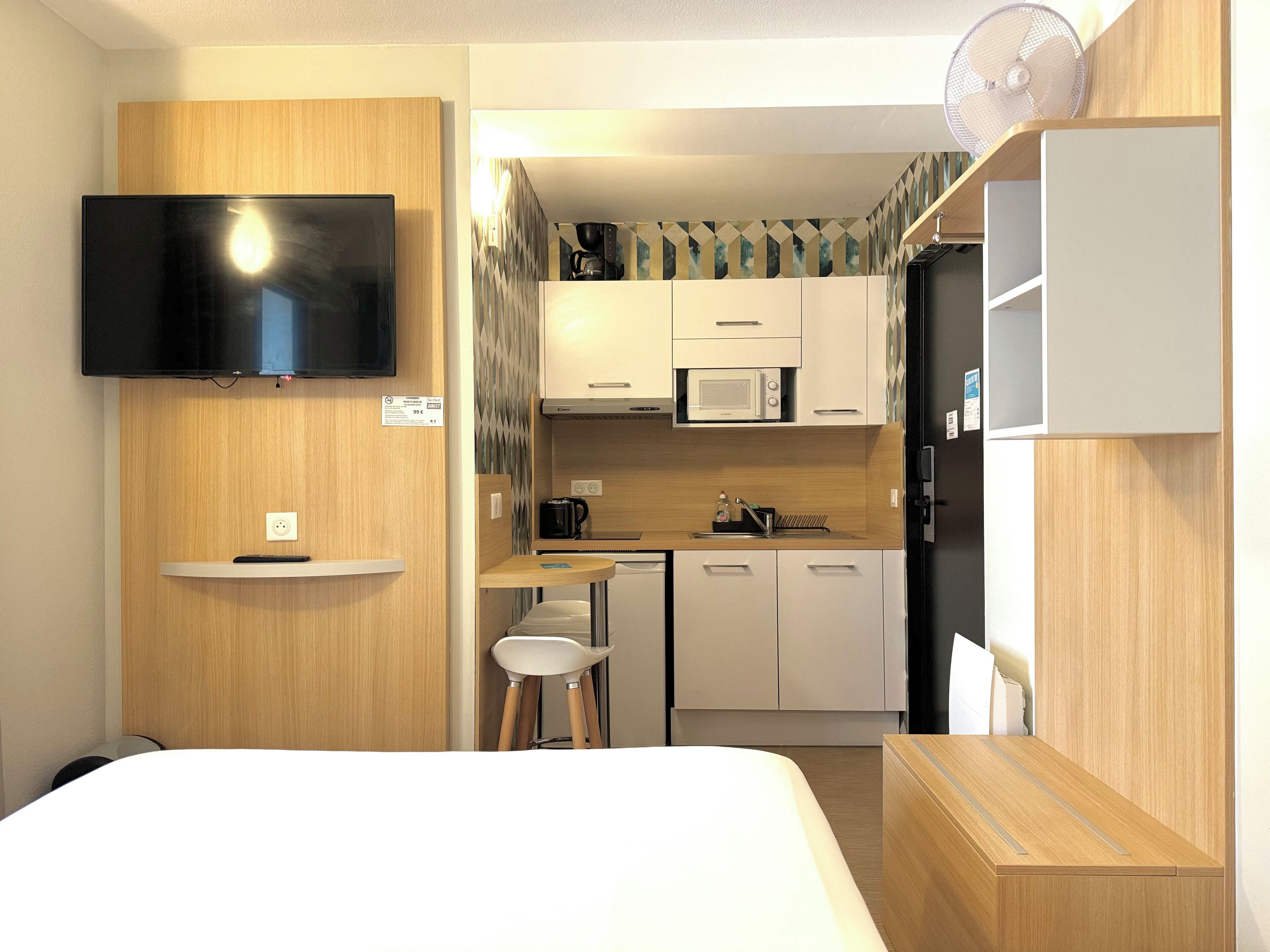 A compact hotel room with a bed, a TV mounted on the wall, a kitchenette with a microwave, a coffee maker, and a small dining area with stools.