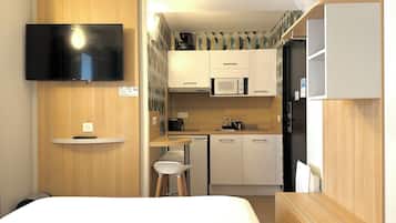 A compact hotel room with a bed, a TV mounted on the wall, a kitchenette with a microwave, a coffee maker, and a small dining area with stools.