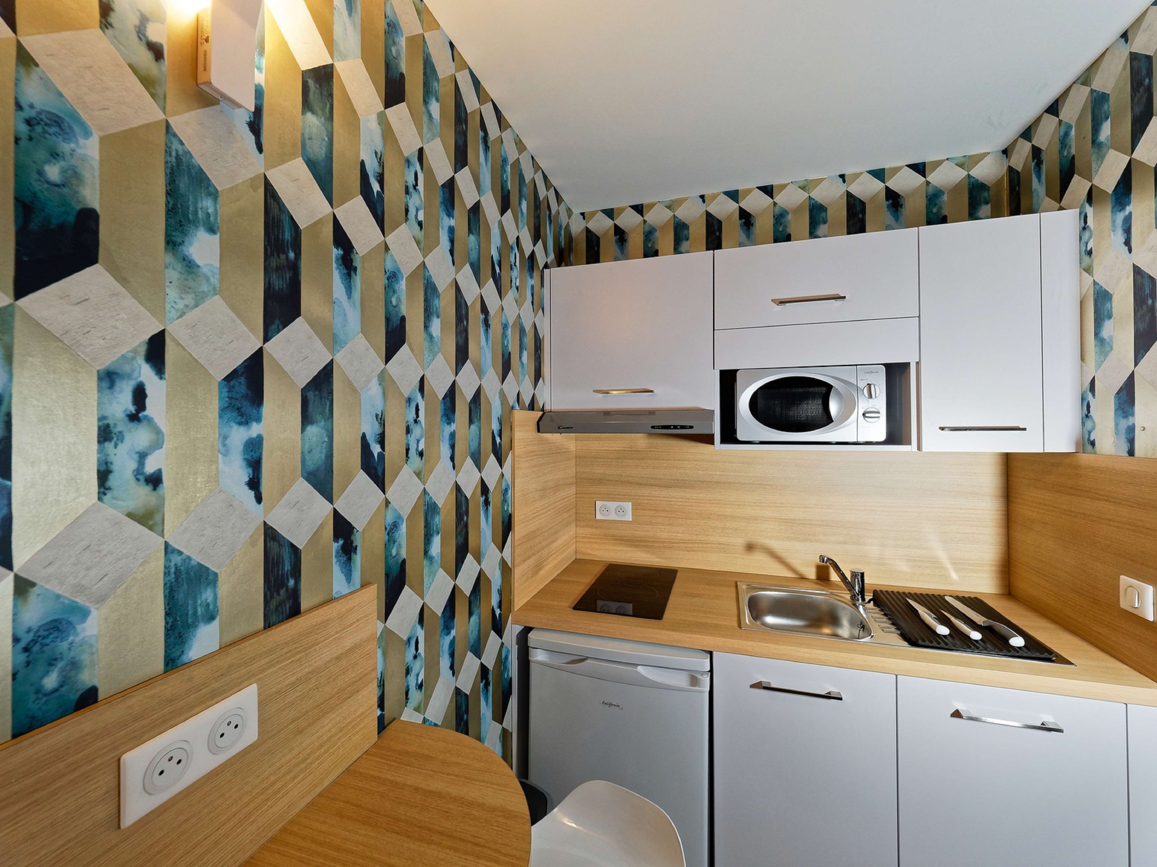 Cocina compacta con microondas, pileta y alacenas, destacando un pared con diseño geométrico.