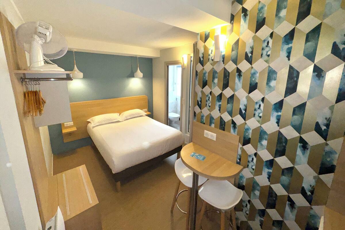 A hotel room with a bed, a small table with two chairs, a wall-mounted fan, and a geometric-patterned wall.