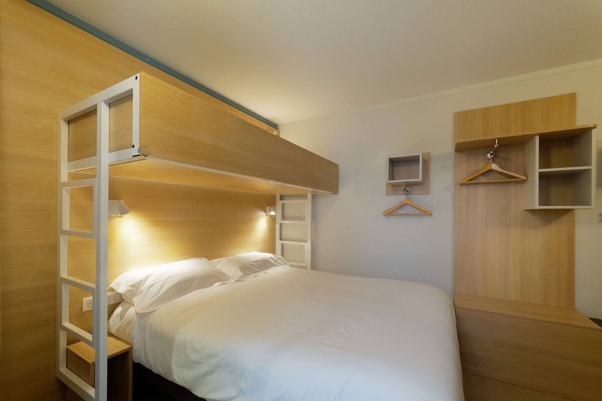 A bunk bed with a wooden headboard and a bedside table. There is a wooden wardrobe with a hanging space and a small shelf.
