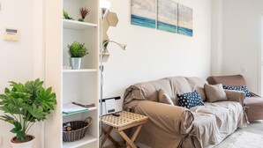 Una sala de estar con un sofá color crema, una mesita de madera, una lámpara de pie y una estantería con plantas.