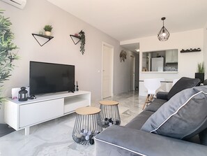 A modern living room with a flat-screen TV, a sofa, a coffee table, and a dining area with a kitchen in the background.