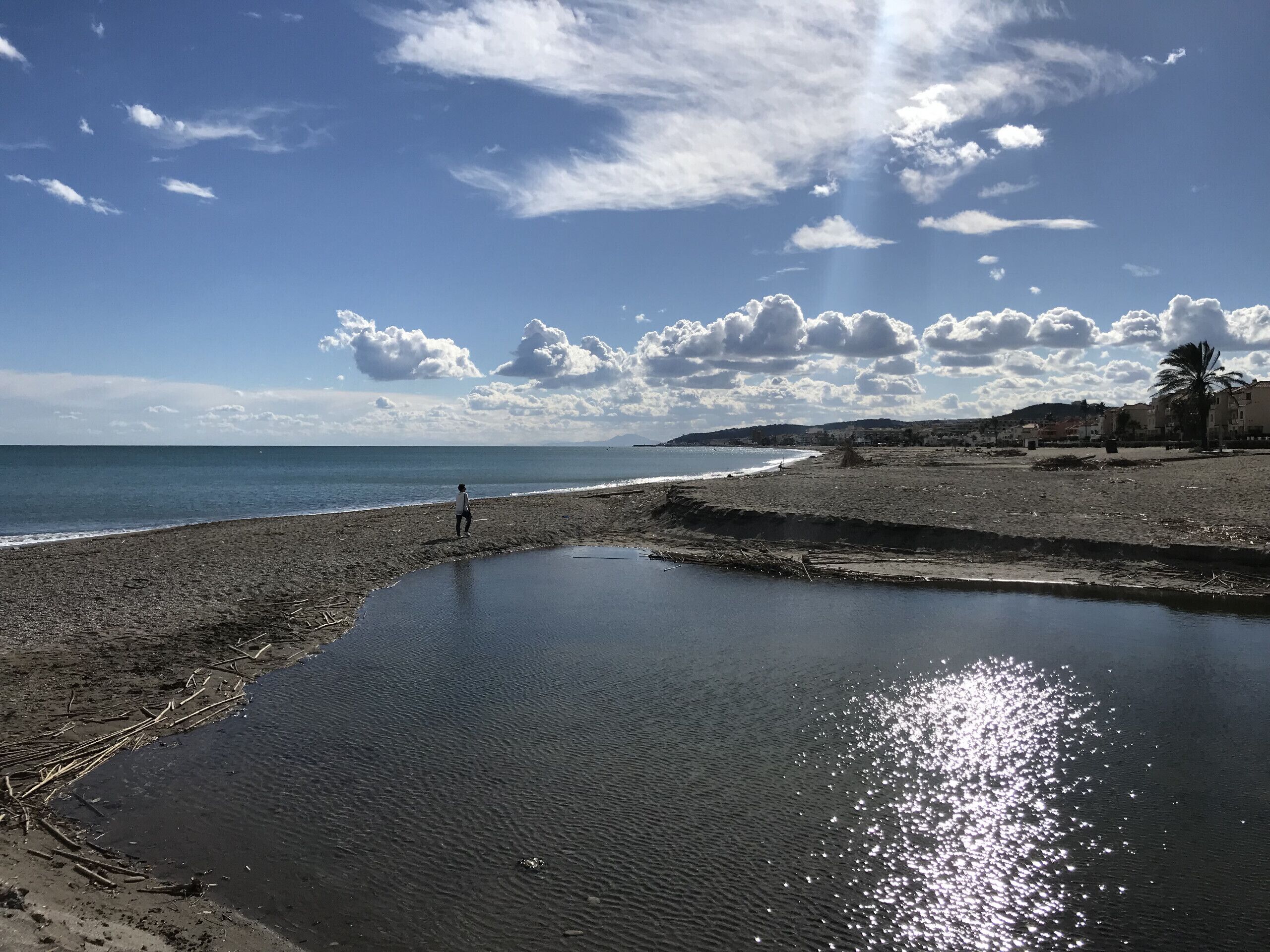 Una playa con una persona caminando por la orilla, un mar tranquilo y un cielo despejado con nubes.