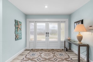 Un pasillo con un tapete estampado, una puerta de cristal con paneles satinados, una decoración con forma de estrella de mar y una lámpara sobre una consola.